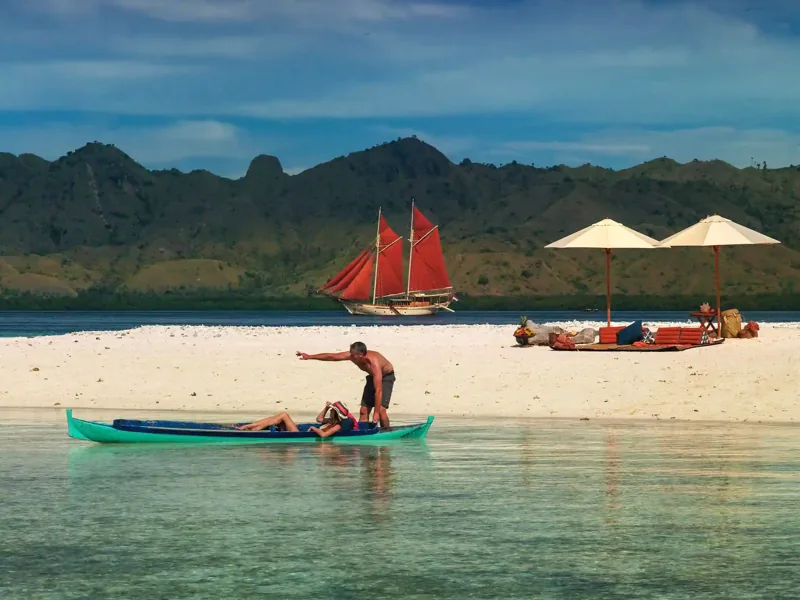 Red-sailed phinisi anchored off a white beach set up for guests in Komodo